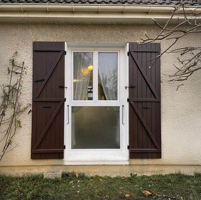 Façade extérieure de maison avec fenêtre à guillotine blanche encadrée de volets battants en bois brun foncé