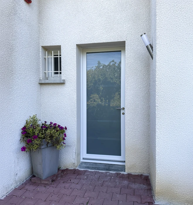 Entrée d'habitation moderne avec porte vitrée à cadre blanc et poignée chromée, mur crépi blanc, fenêtre à barreaudage décoratif, jardinière avec fleurs violettes sur sol pavé en briques rouges disposées en chevrons