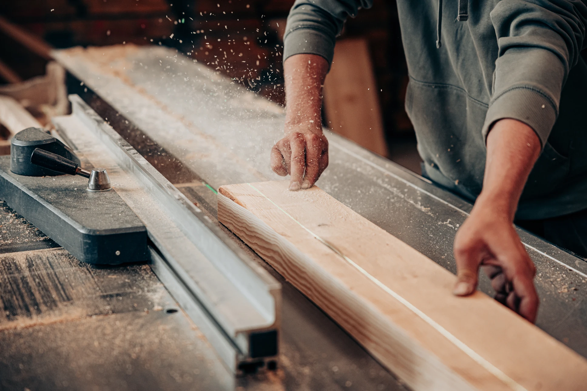 Atelier menuiserie intérieur avec artisan barbu souriant en chemise à carreaux rouge et bleu, posant derrière établi en bois massif